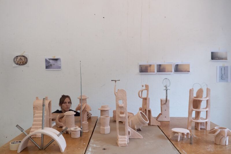 Carolina Fusilier crouching behind a table covered with various abstract clay sculptures.