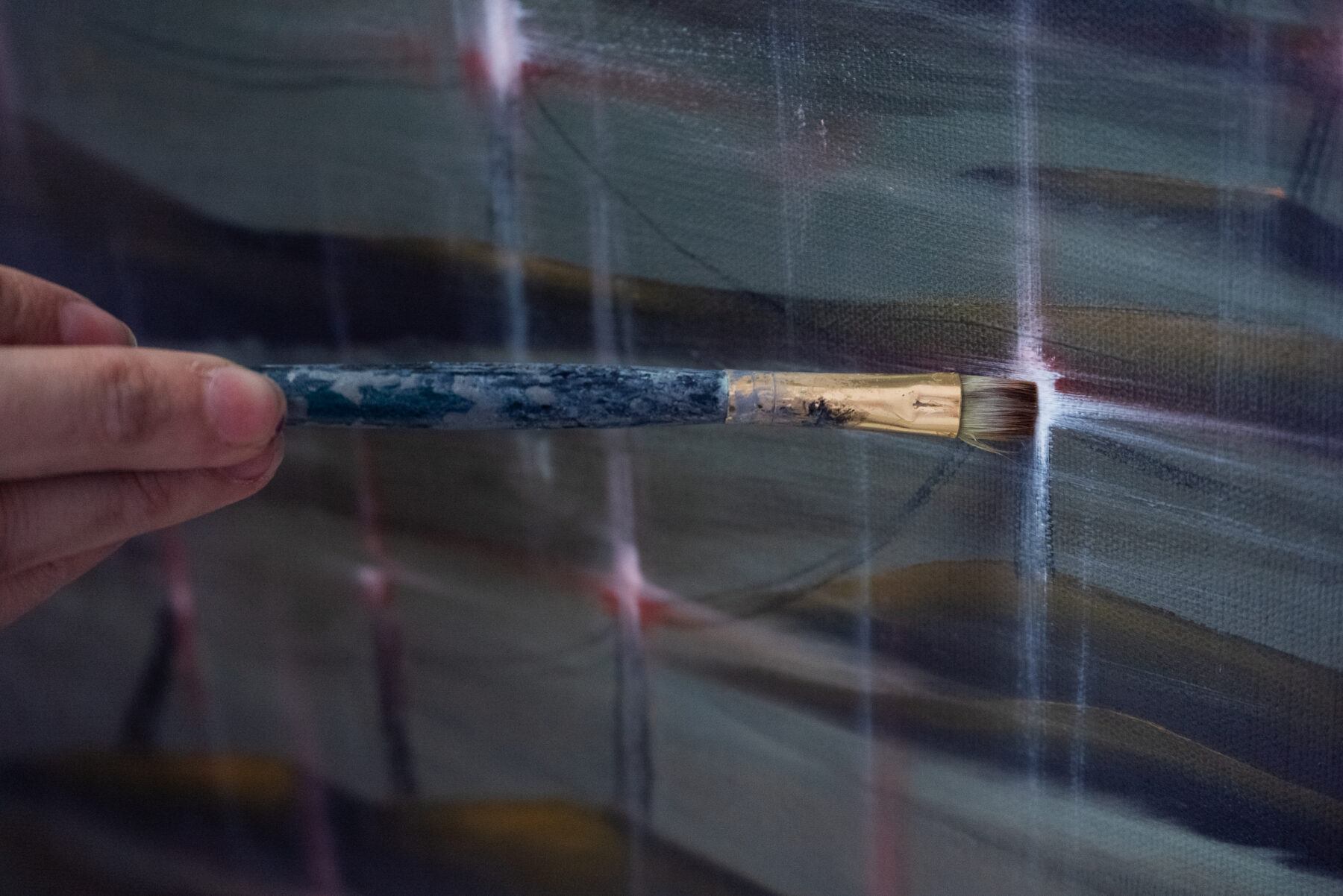 A hand holding a small paintbrush to a canvas, creating horizontal and vertical strokes.