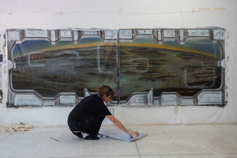 Carolina Fusilier crouched on the floor, working on a large mural-sized canvas featuring futuristic window designs as a border a and an landscape in the background.