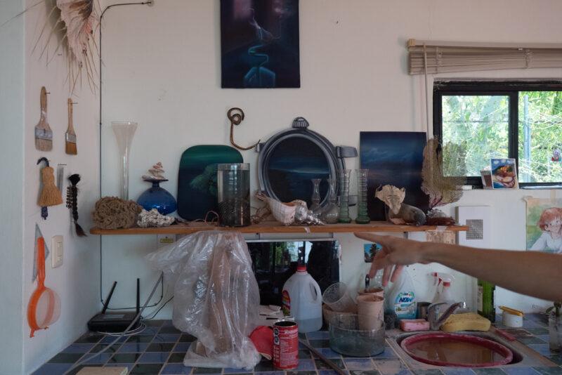 A cluttered shelf with art supplies, seashells, and paintings. A hand points towards the shelf.