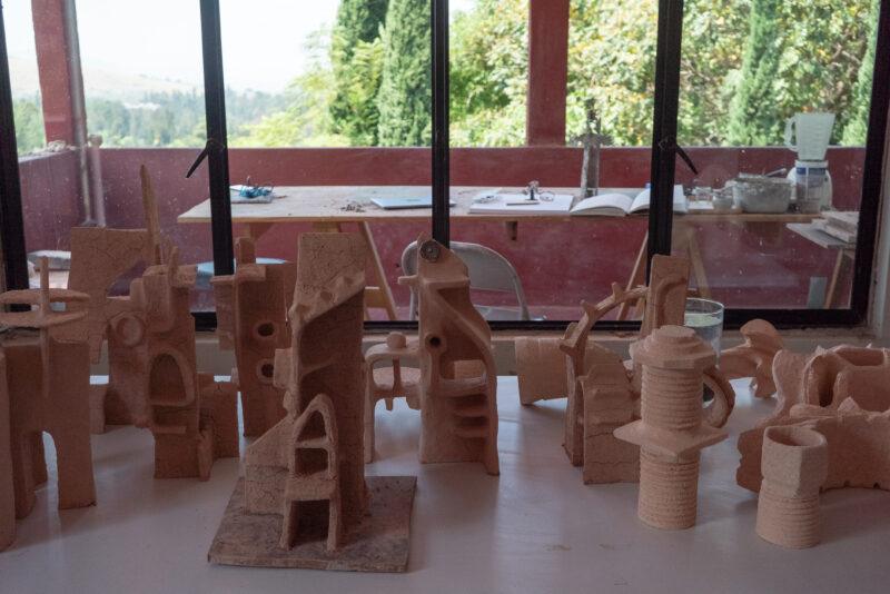 Abstract clay sculptures on a table in front of a large window, with an outdoor scene of trees and a distant view.