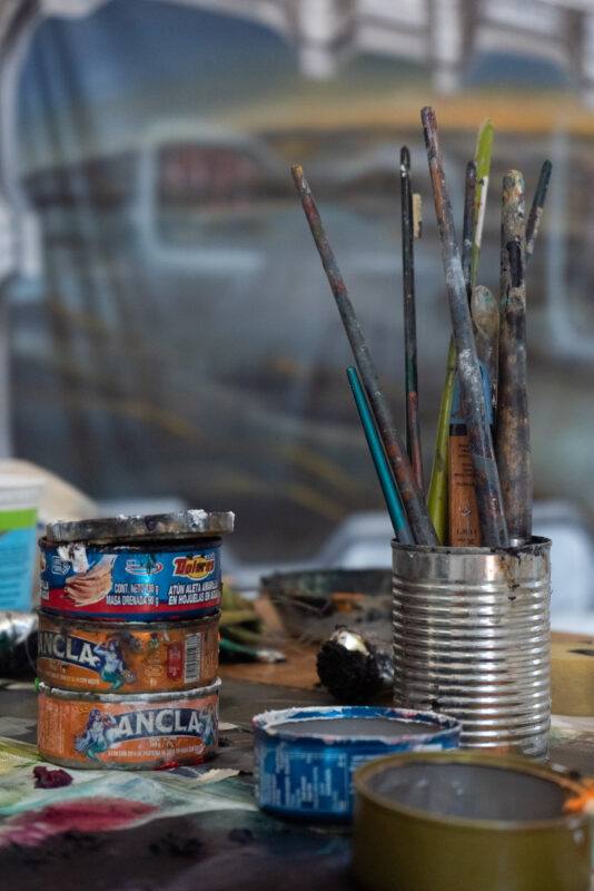 A collection of paintbrushes in a tin and several paint containers are on a table.