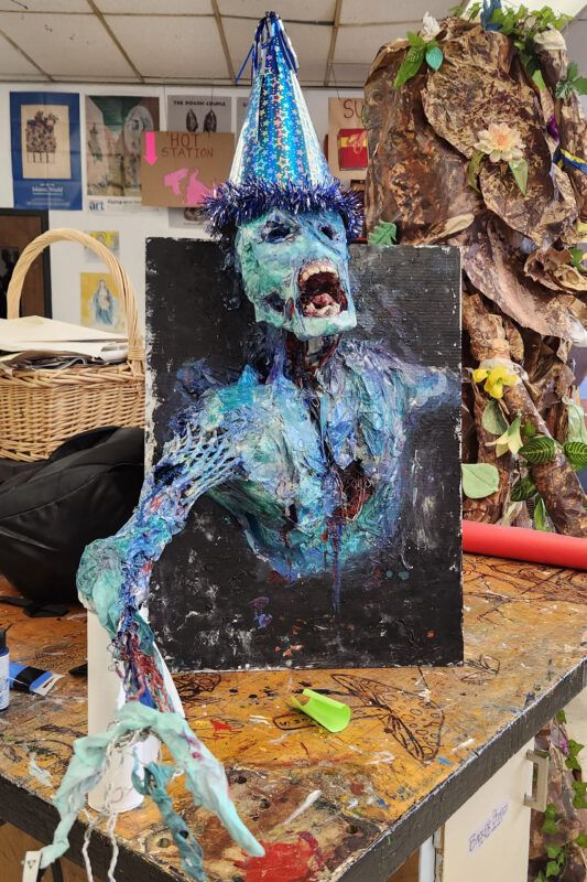 A blue zombie model with a party hat is mounted on a black board, displayed on a cluttered table in an art room.