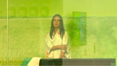 Jade Guanaro Kuriki-Olivo with long hair stands behind a glass panel in a room with green lighting, wearing a white shirt. There are jars on a shelf in the background.