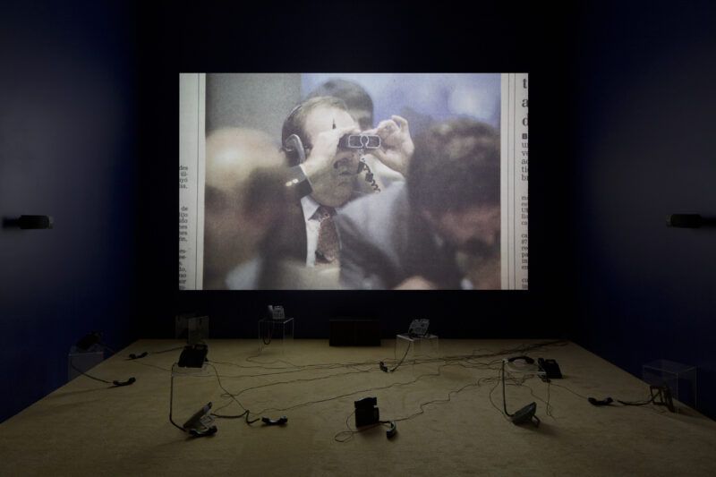 A dimly lit room with a large screen showing a person using binoculars. Wires and speakers are arranged on the floor.