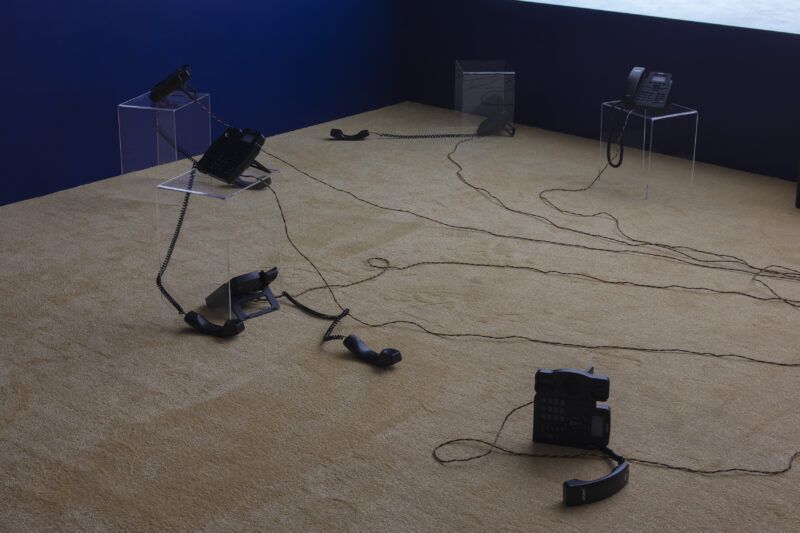 Several corded telephones are placed on a carpeted floor, with tangled wires connecting them. Some phones are on clear stands, while others are lying on the floor in a dimly lit room.