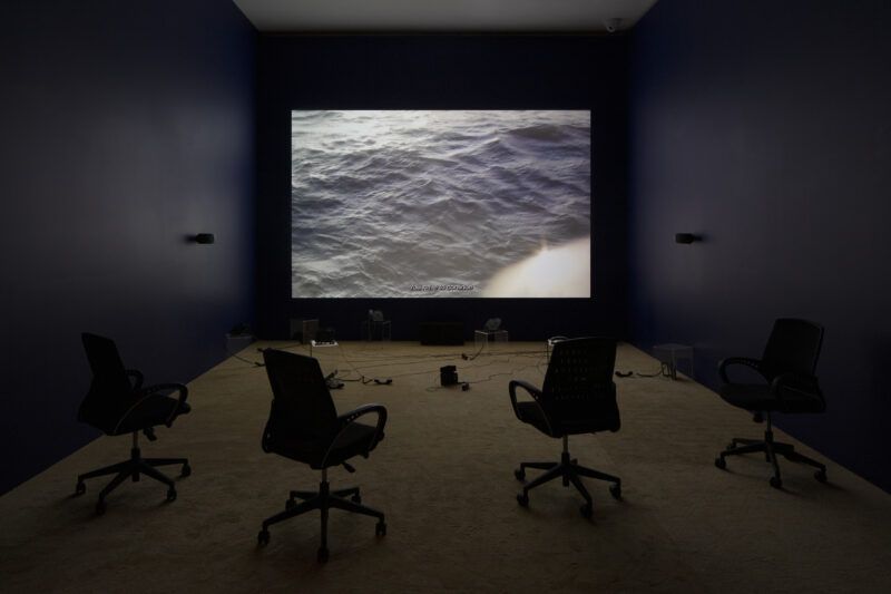 A dimly lit room with four empty office chairs facing a large wall-mounted screen displaying an image of the ocean.