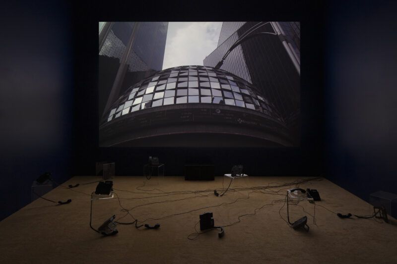 Large screen displays a cityscape with tall buildings. Several black telephones and wires are arranged on the floor in a dimly lit room.