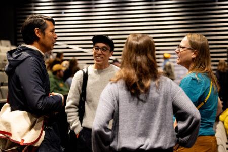 Four people stand in a group talking indoors, facing each other, with striped wall paneling in the background.