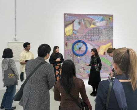 A group of people is gathered in an art gallery, listening to a woman speaking in front of a colorful artwork on the wall.