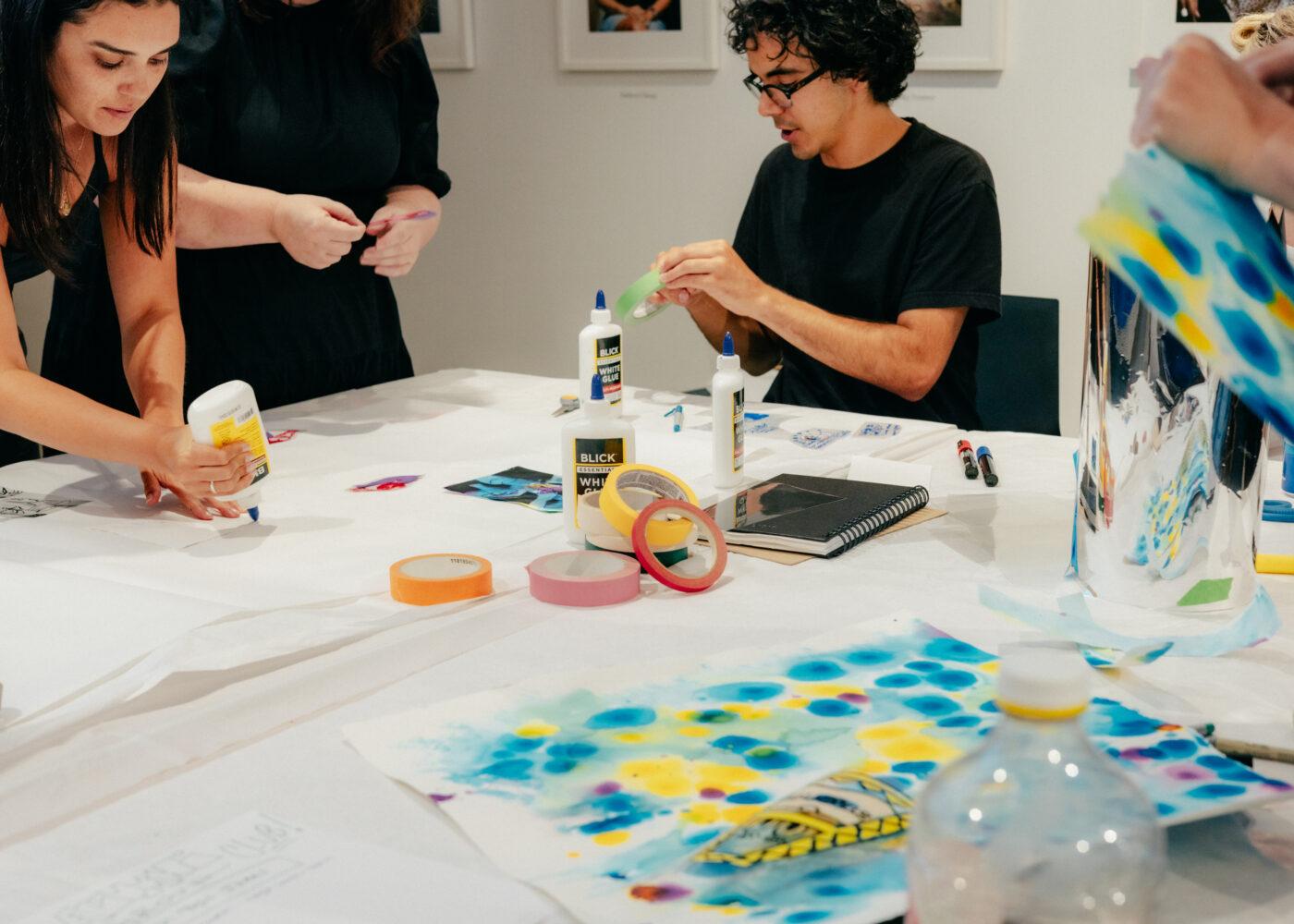 People engaged in an art-making activity at a table, using materials like glue, tape, and colored paper.