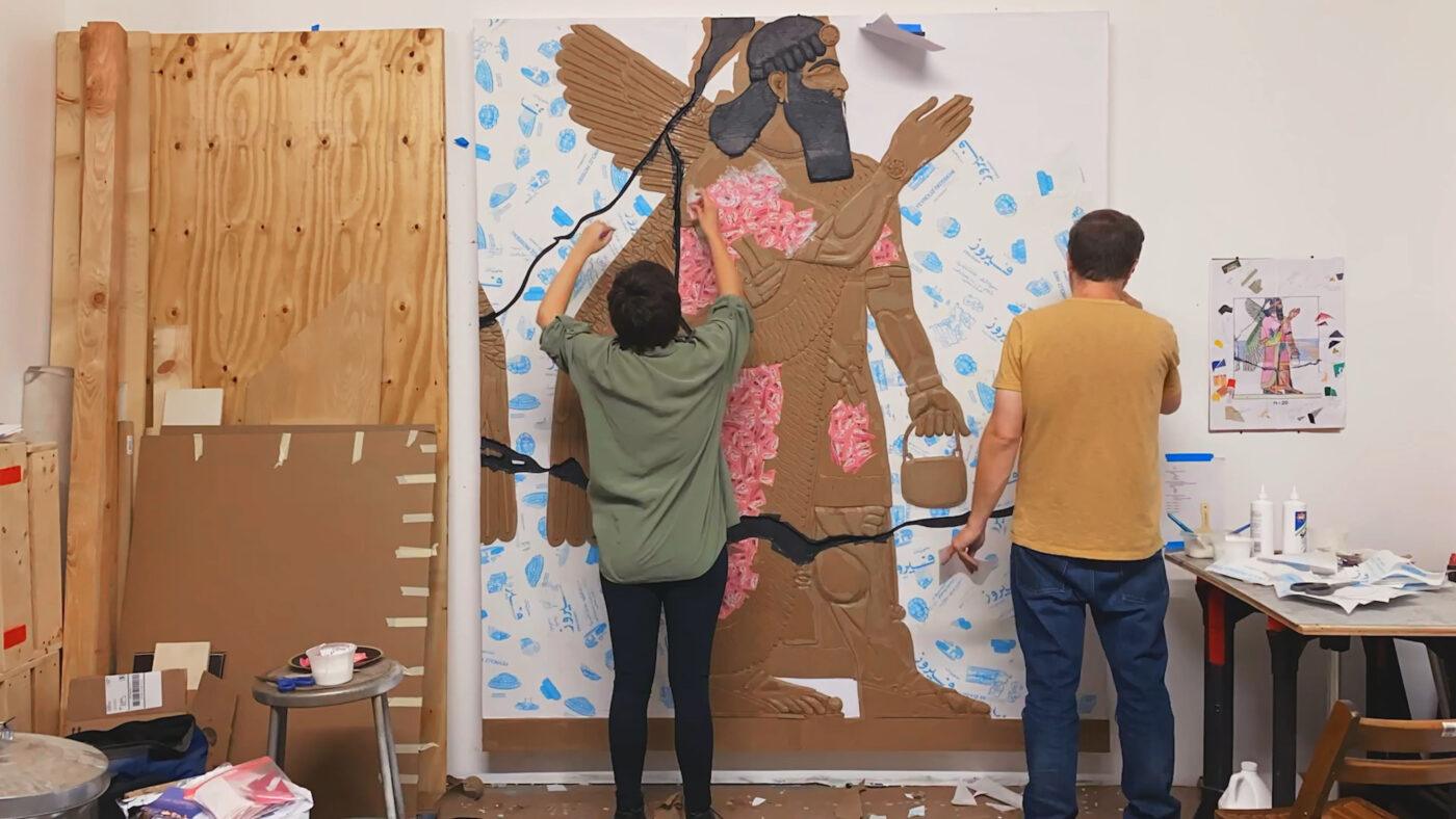 Two people work on a large wall art piece depicting a figure with wings, using paint and mixed materials in a studio space.