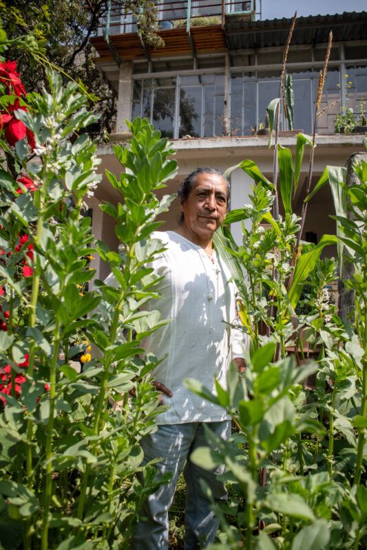 Fernando Palma Rodríguez stands in a lush garden filled with tall green plants and red flowers, in front of a multi-story house with exposed wooden balconies.