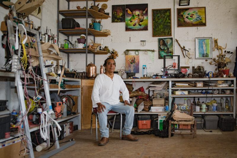 A person sitting calmly on a stool in a cluttered workshop surrounded by shelves filled with tools, containers, and various objects. The background wall is decorated with multiple artwork pieces and photographs.