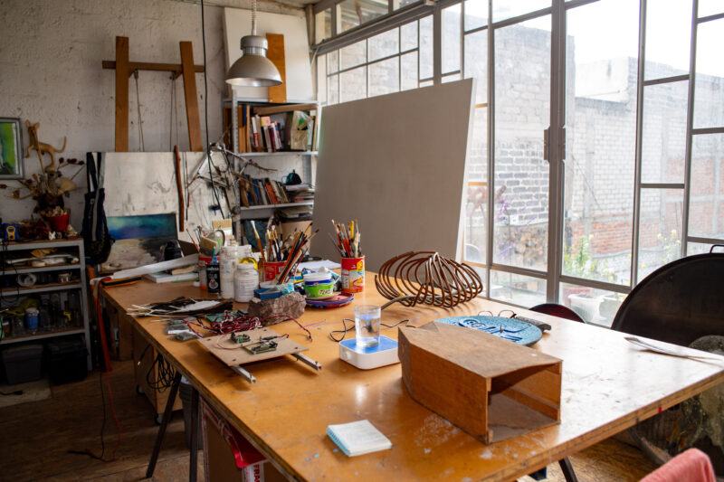 An artist's studio filled with various art supplies, unfinished canvases, and tools, illuminated by natural light from large windows.