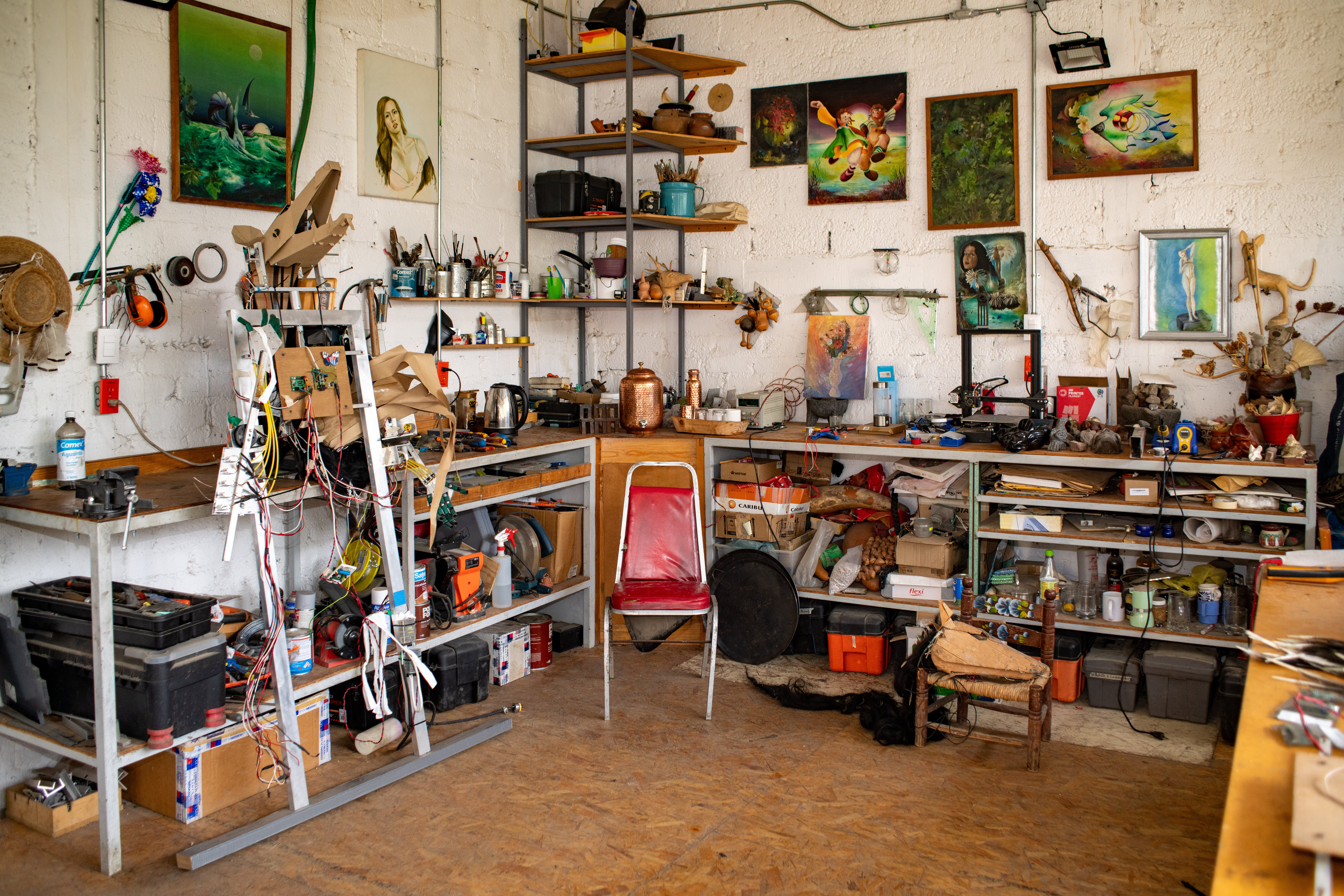 Interior of a cluttered workshop with various tools, equipment, and artwork on the walls and shelves. A red chair sits in the center.