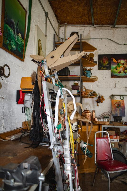 Interior of a cluttered workshop filled with various tools, artworks, and a robotic assembly. The space is adorned with paintings, sculptures, and mechanical parts.