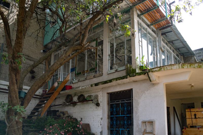 Exterior of a rustic building, featuring a staircase leading to a second-floor balcony shaded by trees, and a blue door with ornamental designs on the groudn level.