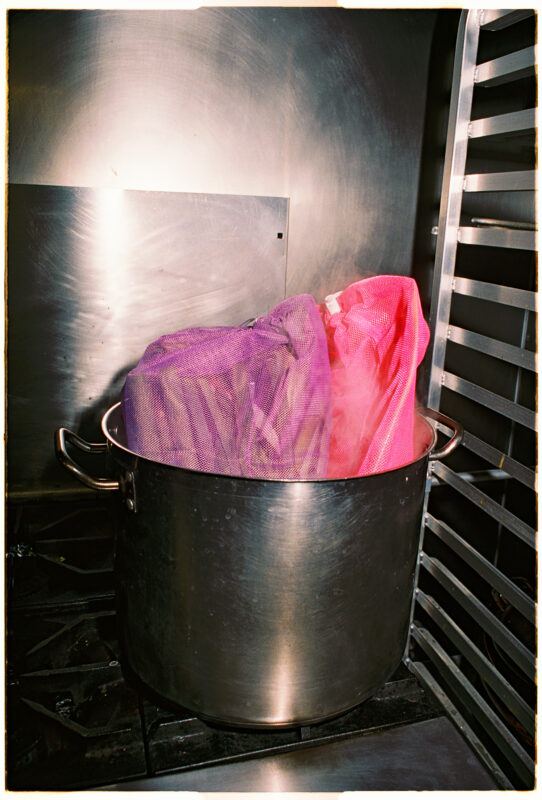 Two mesh bags, one pink and one purple, are placed inside a large metal pot on a stove.