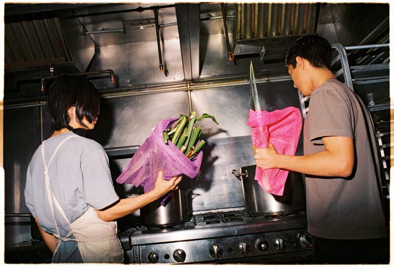 Two people in an industrial kitchen, each holding purple net bags with leafy green vegetables, standing in front of a stove with two pots.