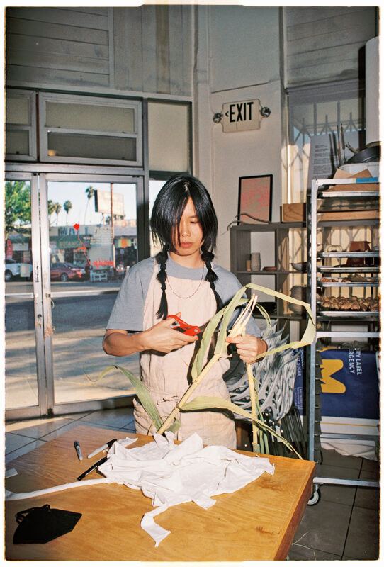 Ami Lien in an apron using scissors to cut green leaves at a wooden table, inside a room with large windows and an exit sign.