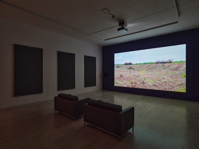 A dimly lit room features two couches facing a large screen displaying an outdoor scene with vehicles in an open field.