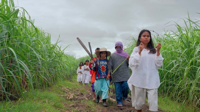 A group of children, walk through a lush green field under a cloudy sky.