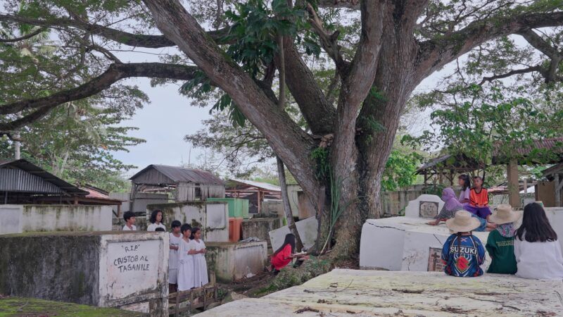 A group of people, including children in white attire, gather in a cemetery under a large tree.