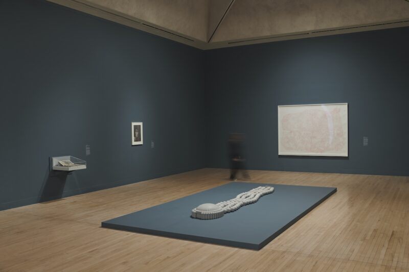 Art gallery with dark grey walls, featuring a sculpture on a low platform, two framed artworks, and book encased in a glass case attached to the wall. A person is captured in motion walking past across the wooden floor.