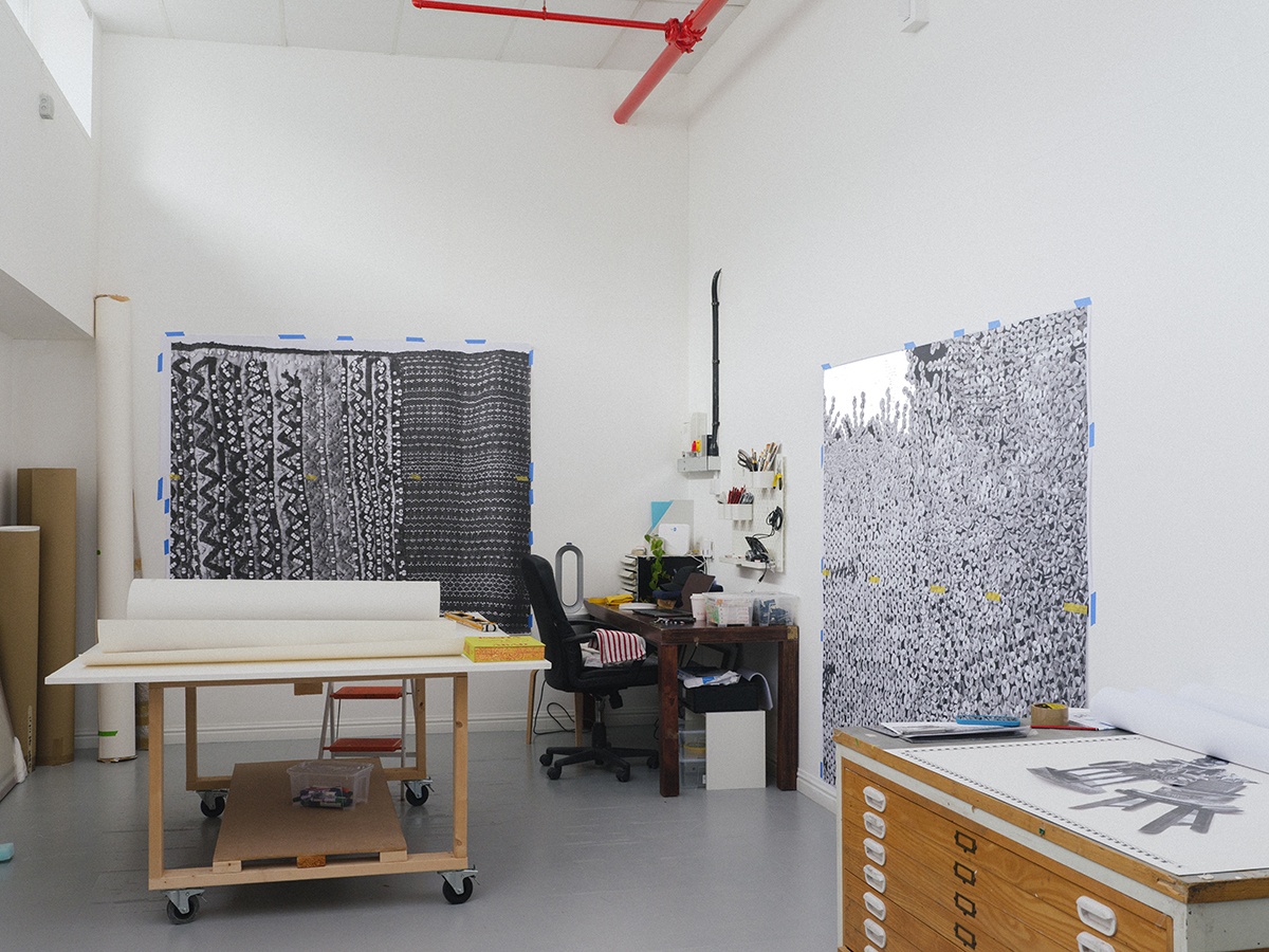 Art studio with large black and white patterned artworks on walls, work table with paper rolls, a desk with chair, and storage drawers.