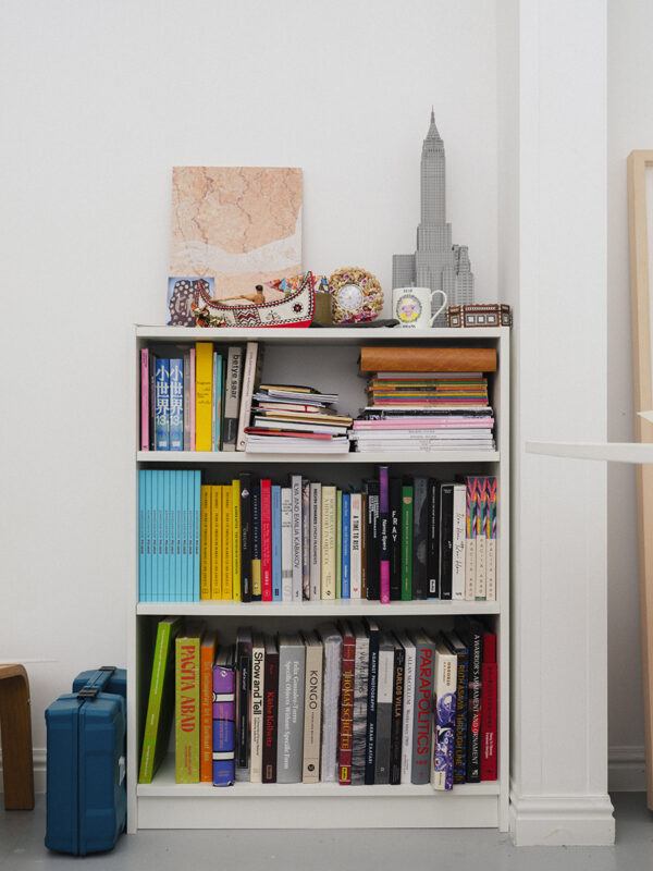 A white bookshelf filled with various books and magazines. On top, there's a model of the Empire State Building, a decorative box, and a framed artwork. A blue case is on the floor beside it.