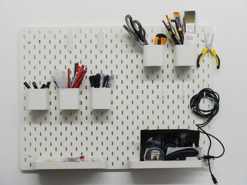 White pegboard mounted on a wall, holding containers with pens, scissors, brushes, pliers, cables, and a shelf with tape and other stationery items.