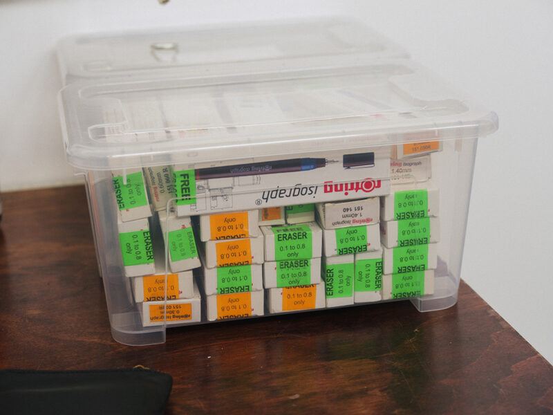 A transparent plastic container on a wooden surfaced filled with numerous boxes labeled Eraser in green and orange stickers, along with a box of pens.