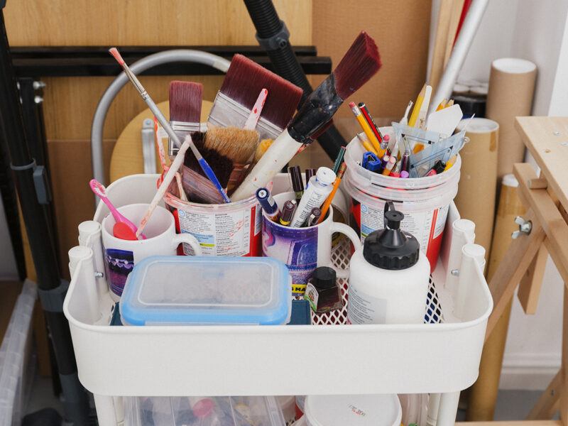 A cart holds a variety of art supplies, including paintbrushes, pencils, markers, and containers, against a studio backdrop.