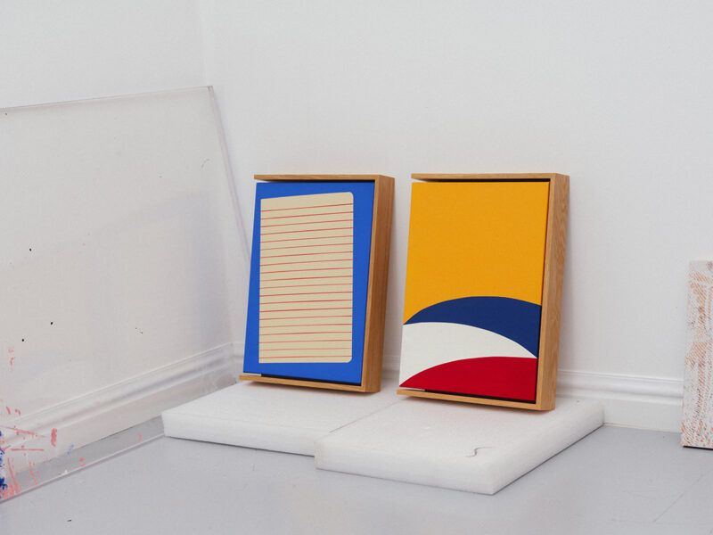 Two wooden frames lean against a white wall. One has a painted lined paper design; the other displays yellow, blue, and red abstract shapes. They are placed on rectangular white blocks.