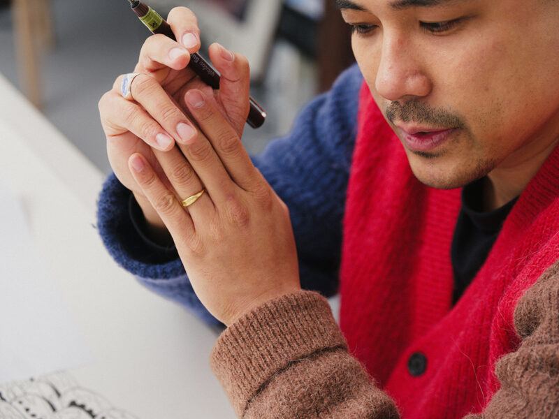 Pio Abad wearing a multicolored sweater holds a pen while focused on drawing.