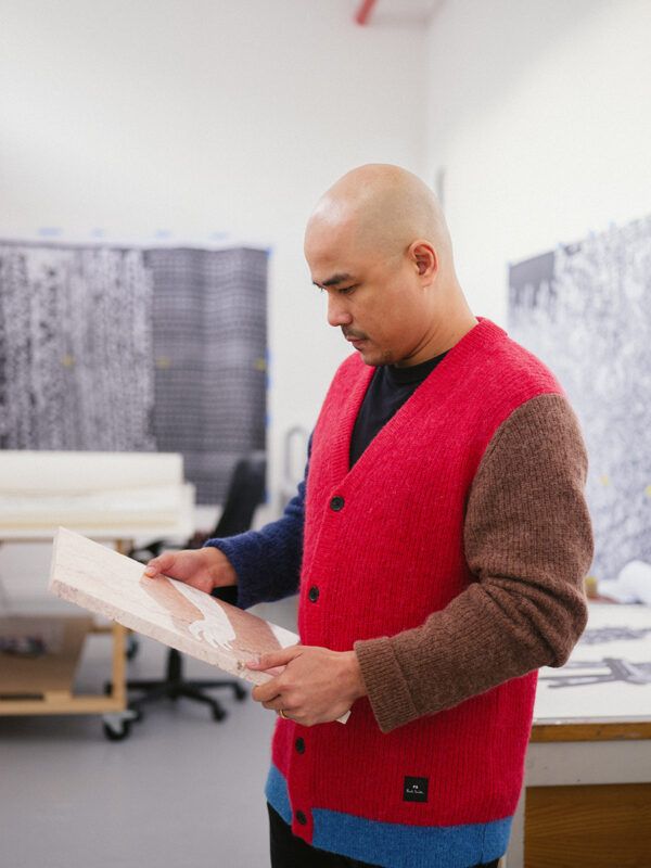 Pio Abad in a colorful sweater examines a piece of artwork in a studio filled with canvases and materials.