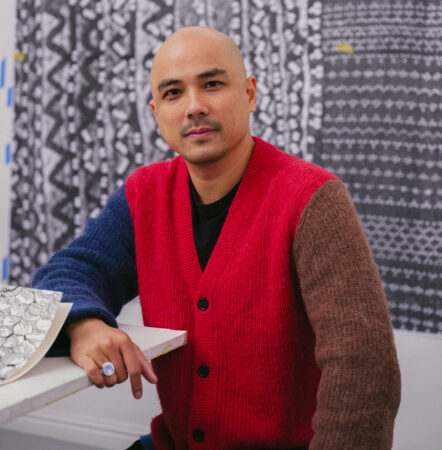 Pio Abad in a multicolored cardigan sits in front of black and white pattern on the wall, looking at the camera.