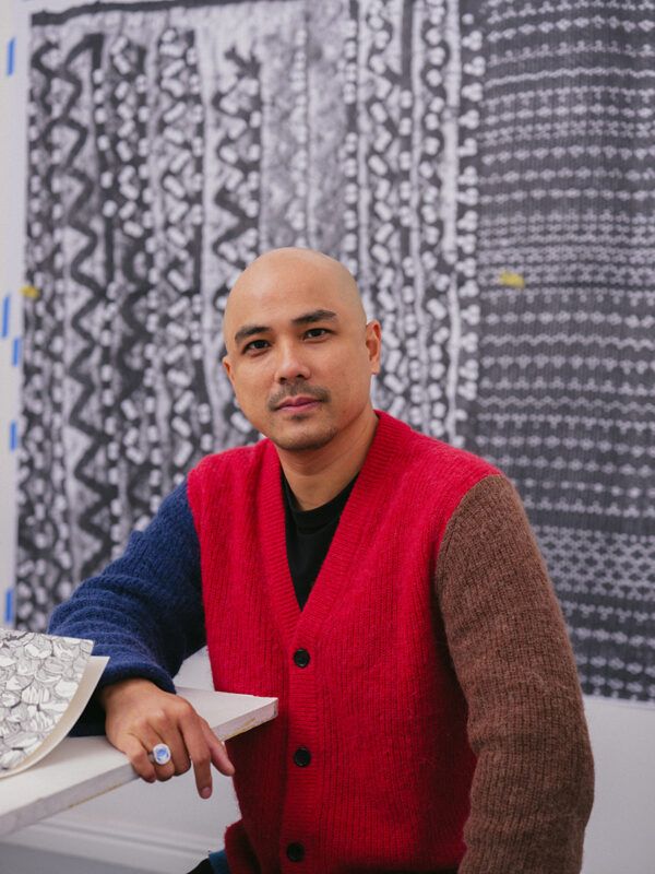 Pio Abad in a multicolored cardigan sits in front of black and white pattern on the wall, looking at the camera.