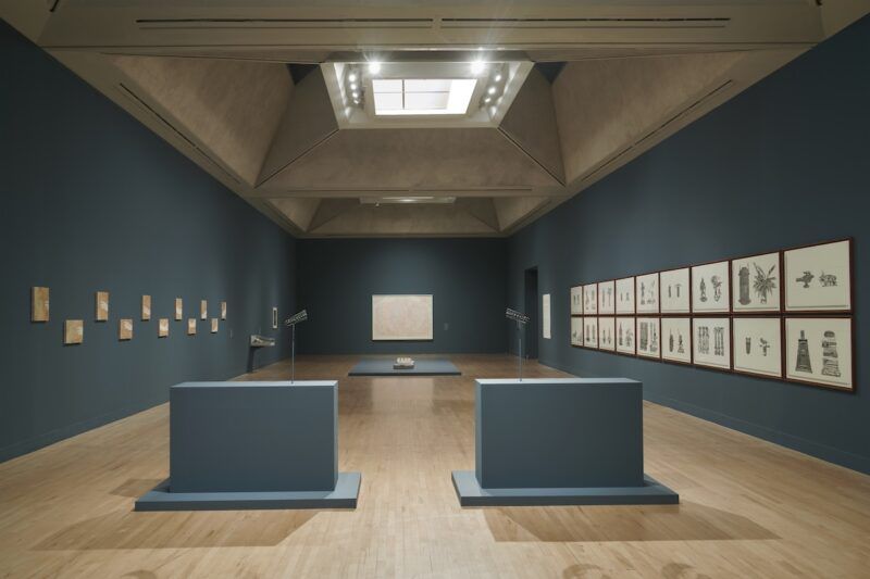 Art gallery with dark grey walls featuring framed artworks. A large ceiling light illuminates the room, with two display stands and a central sculpture on the floor.