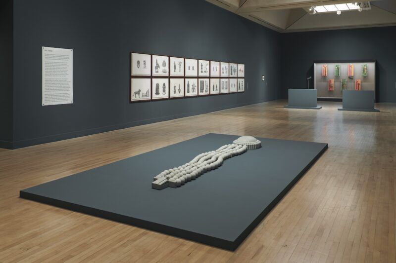 Gallery featuring a large white beaded sculpture displayed on a grey platform on the floor. Throughout the space framed illustrations are on the walls, and colorful elongated objects are in a display case in the rear of the room.