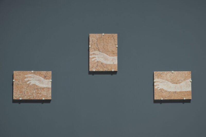 Three artworks on a dark wall display hands reaching out, with each piece featuring a marble-like texture in the background.