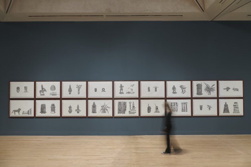 A person walks past a series of framed illustrations on a dark grey wall in a gallery.