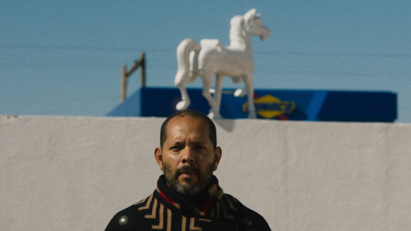 Guadalupe Maravilla stands in front of a white wall with a white horse statue on top, a blue building in the background.