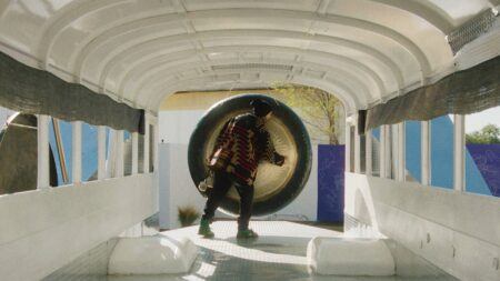 Guadalupe Maravilla with a patterned jacket and hat stands inside a white empty bus, reaching towards a large metallic gong.