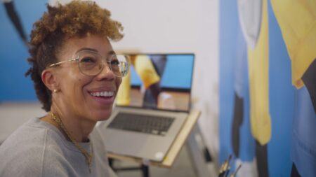 Amy Sherald with curly hair and glasses smiling, seated next to an open laptop.