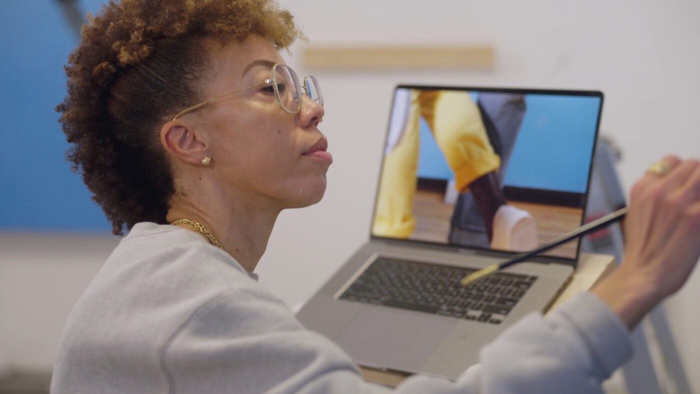 Amy Sherald with glasses and curly hair painting while looking at a laptop displaying an image of a person wearing yellow pants.