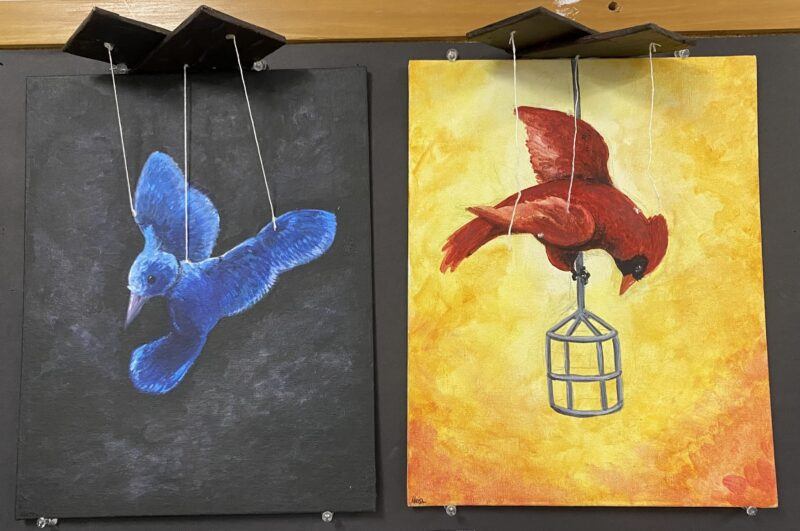 Two paintings: left shows a blue bird on a dark background, right shows a red bird with a cage on a yellow-orange background. Both are suspended with strings.