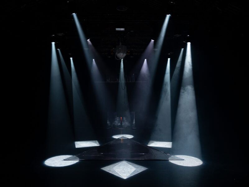 A dark stage with multiple spotlights illuminating triangular and diamond-shaped patterns on the floor.