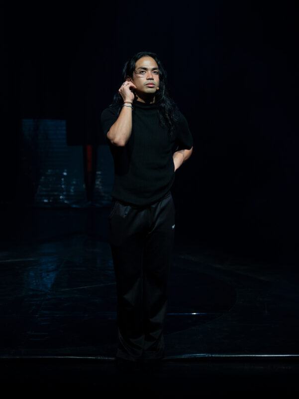 Person with long hair and black outfit stands on a dimly lit stage with one hand on their hip and the other hand raised to their face.